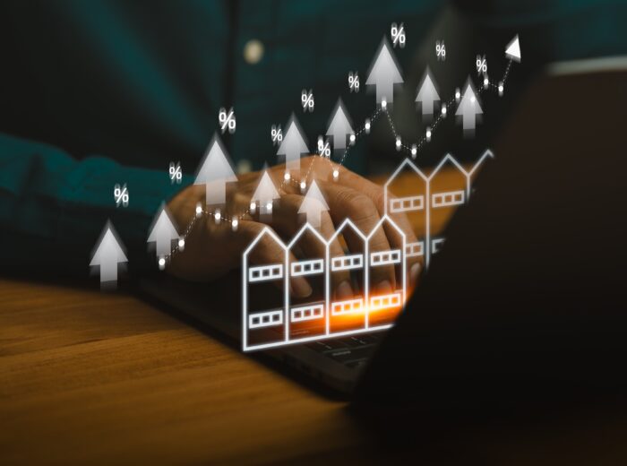 Real estate market growth concept showing person using laptop with rising commercial building icons, percentage symbols, and upward arrows, representing property investment, shophouse, housing trends.