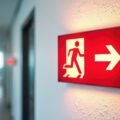 Illuminated red emergency exit sign mounted on office wall showing running man icon and right arrow. Provides clear guidance for evacuation routes and, safety compliance in modern building hallways.