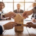 Business team building house model from wooden blocks in meeting, concept of teamwork, real estate planning, mortgage, insurance and investment strategy.