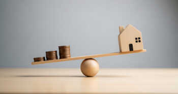 A wooden seesaw balances stacks of coins against a small wooden house, illustrating financial concepts.